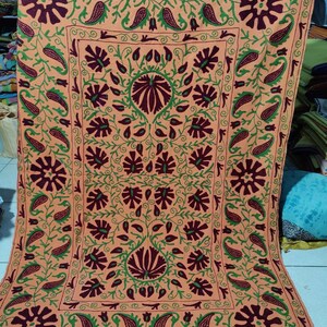 May include: A large, square, peach-coloured throw pillow with intricate embroidery in shades of green and maroon. The design features a repeating pattern of stylised flowers and paisley shapes.