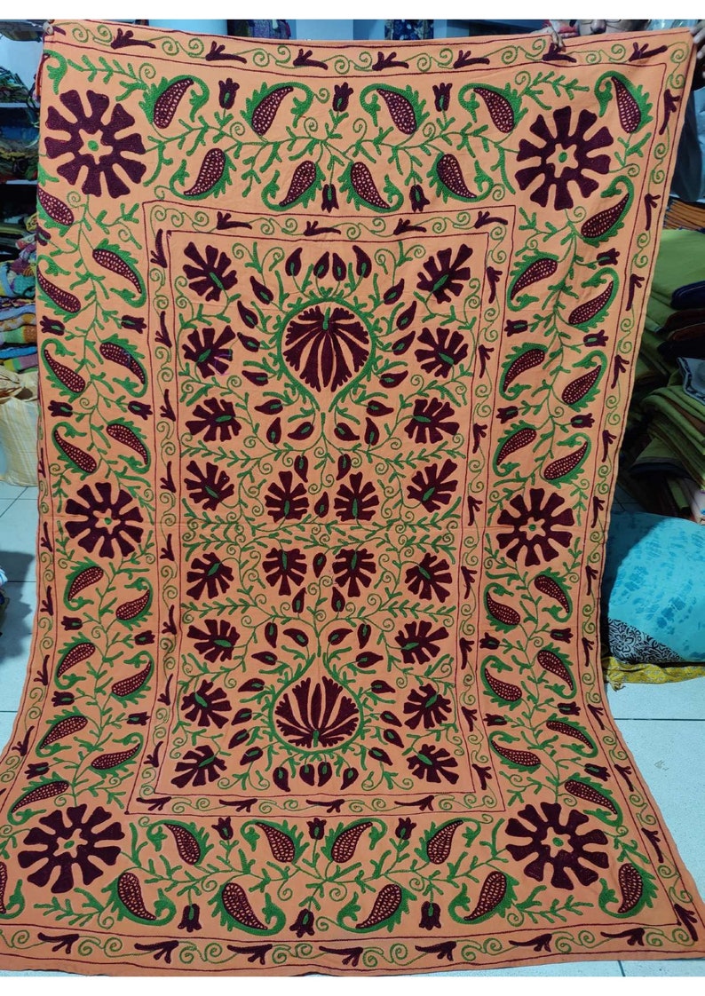 May include: A large, square, peach-coloured throw pillow with intricate embroidery in shades of green and maroon. The design features a repeating pattern of stylised flowers and paisley shapes.