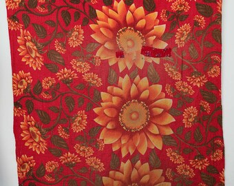 May include: A rectangular textile featuring a vibrant red background with a repeating pattern of large orange and yellow sunflowers, smaller orange blossoms, and dark green leaves. The fabric displays visible fine stitching throughout.