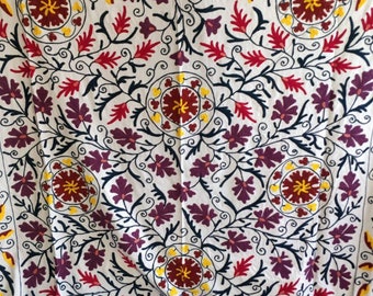 May include: White fabric with a repeating pattern of embroidered flowers in shades of red, yellow, and purple. The flowers are outlined in black thread and have a traditional design.