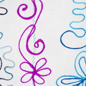 May include: White fabric with embroidered floral designs in pink, blue, and black. The design features swirls, flowers, and leaves.