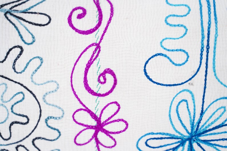 May include: White fabric with embroidered floral designs in pink, blue, and black. The design features swirls, flowers, and leaves.