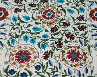 May include: White fabric with intricate embroidery featuring a floral pattern in shades of blue, green, red, and orange. The design includes stylised flowers, leaves, and swirls.
