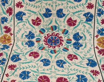 May include: A cream-colored fabric square with a repeating pattern of blue and red flowers embroidered on it. The flowers are surrounded by green leaves and vines.