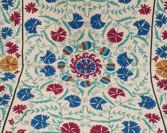 May include: A cream-colored fabric square with a repeating pattern of blue and red flowers. The flowers are embroidered with a green stem and leaves. The center of the fabric features a larger, more intricate design with a blue and red flower.