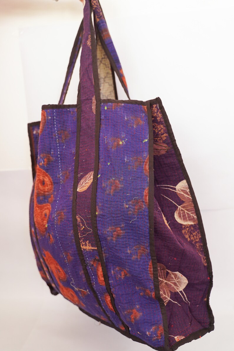 Puede incluir: Un bolso tote grande, morado y marr&oacute;n con un dise&ntilde;o de patchwork. El bolso tiene un fondo ancho y plano y dos asas largas.