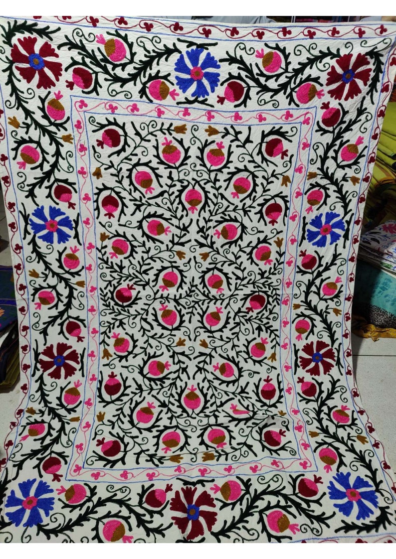 May include: A white cotton fabric with intricate embroidery featuring a floral design with pink, brown, and blue flowers. The embroidery is done in black thread and creates a detailed pattern.