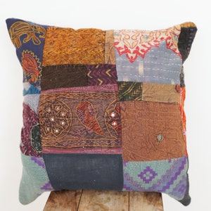 Puede incluir: Una funda de cojín de patchwork con una variedad de colores y texturas, incluyendo marrón, azul, morado y naranja. La funda está hecha de diferentes telas y tiene intrincados detalles de bordado.