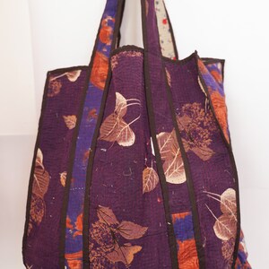 Puede incluir: Una gran bolsa de mano de color morado con correas marrones y estampado floral. La bolsa est&aacute; hecha de tela acolchada y tiene un dise&ntilde;o &uacute;nico de patchwork.