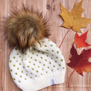 May include: White knit beanie with a green pom pom and a brown faux fur pom pom. The beanie has a small white tag that says "Miniko".
