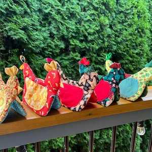 May include: Five fabric chicken-shaped ornaments arranged in a row. Each chicken features a unique combination of colourful patterned cotton fabrics, including red, green, yellow, and gingerbread-themed prints, with small fabric loops at the top.