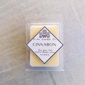 May include: A rectangular soy wax tart in a clear plastic container. The label reads "Cinnabon" with the text "Soy Wax Tart" and "1 OZ / 28 GRAMS". The label also features a dreamcatcher design and the text "Tribe Candle Co."