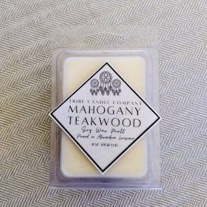 May include: A soy wax melt in a clear plastic container. The label reads "Mahogany Teakwood" and "Tribe Candle Company." The melt is 1 oz (28 grams). The label has a diamond shape with dream catchers.