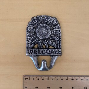 K&ouml;nnte beinhalten: Ein silberfarbenes Willkommensschild aus Metallguss mit Sonnenblumenmuster. Das Schild tr&auml;gt das Wort "WELCOME" am unteren Rand, darunter ein Haken. Ein Lineal liegt neben dem Schild.
