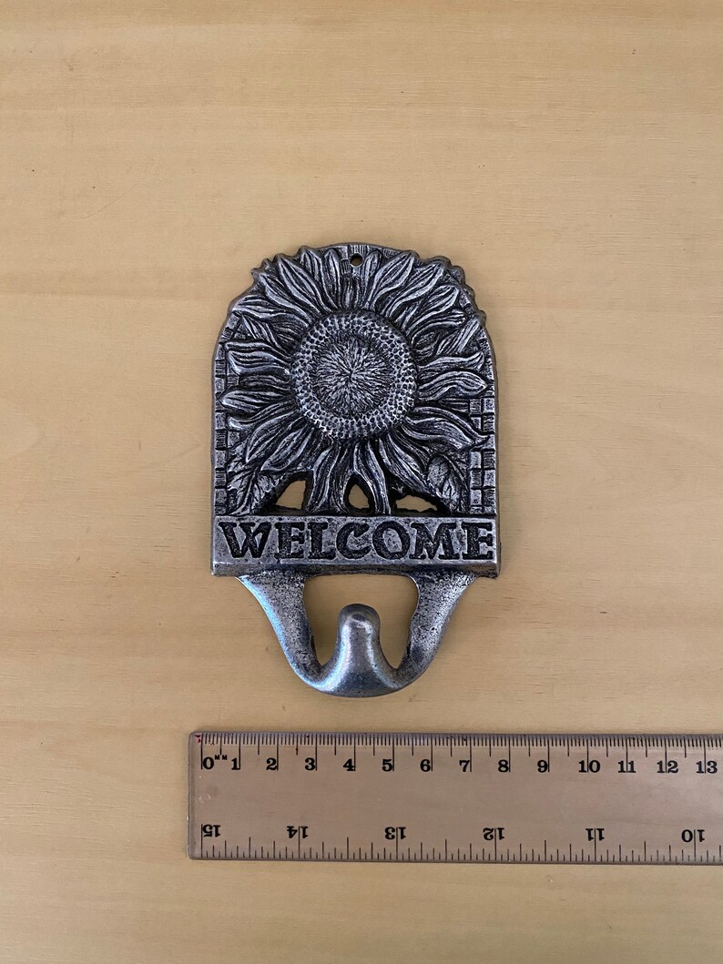 K&ouml;nnte beinhalten: Ein silberfarbenes Willkommensschild aus Metallguss mit Sonnenblumenmuster. Das Schild tr&auml;gt das Wort "WELCOME" am unteren Rand, darunter ein Haken. Ein Lineal liegt neben dem Schild.