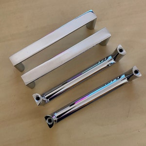 May include: Four polished chrome cabinet pulls on a light wood surface. Two are rectangular bar pulls, and two are tubular with decorative ends. The hardware is reflective, showing the surrounding environment.