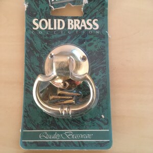May include: A solid brass door pull handle on a green and white card. The handle is a shiny gold colour with a circular base and a curved handle. The card reads "SOLID BRASS COLLECTION" and "Quality Brassware".
