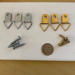 May include: A selection of picture hanging hardware, including silver and gold-coloured D-rings, screws, and a coin. The D-rings have a triangular base and a hole for hanging. The screws are silver and gold. The coin is silver.