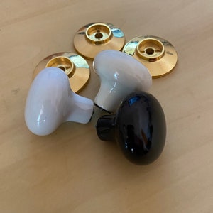 May include: A selection of cabinet knobs and backplates. The knobs are white and black, with a glossy finish. The backplates are gold-coloured and circular. The knobs are oval-shaped and appear to be made of ceramic.