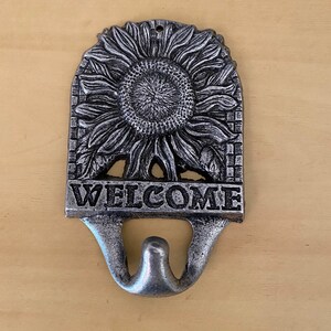 K&ouml;nnte beinhalten: Ein Willkommensschild aus Gussmetall mit Sonnenblumenmuster. Das Schild ist silberfarben und tr&auml;gt das Wort "WELCOME" in fetten Buchstaben. Unter dem Wort "WELCOME" befindet sich ein Haken.