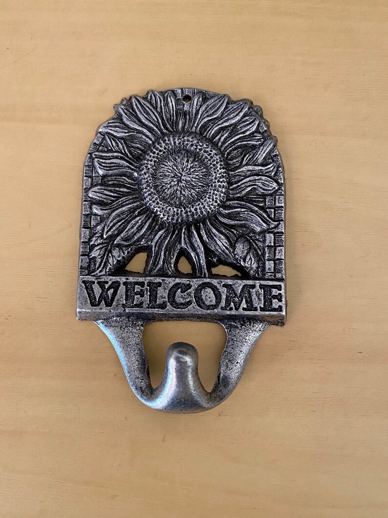 K&ouml;nnte beinhalten: Ein Willkommensschild aus Gussmetall mit Sonnenblumenmuster. Das Schild ist silberfarben und tr&auml;gt das Wort "WELCOME" in fetten Buchstaben. Unter dem Wort "WELCOME" befindet sich ein Haken.