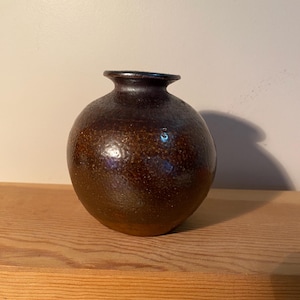 May include: A small, round, brown ceramic vase with a narrow neck. The vase has a textured surface and a glossy finish, with variations in colour from dark brown to a lighter, speckled brown. The vase sits on a wooden surface.