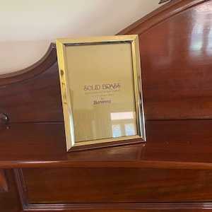May include: A rectangular, polished brass photo frame. The frame is 7" x 7" (18cm x 18cm) and features the text "SOLID BRASS" and "hand polished & lacquer coated". The frame is by Bowen.