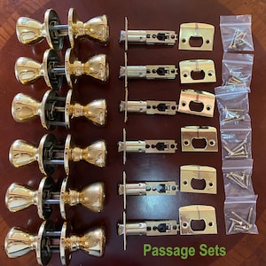 May include: A selection of polished brass door passage sets. The sets include doorknobs, latches, strike plates, and screws, arranged on a wooden surface. The text "Passage Sets" is visible.