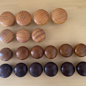 May include: A selection of wooden cabinet knobs in shades of brown, arranged in rows. The knobs vary in colour from light to dark, with visible wood grain patterns. The knobs are round and appear to be made of solid wood.