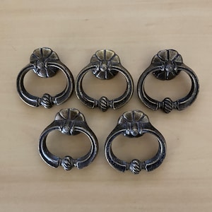 May include: Five antique brass-finished metal ring pulls arranged in a group. Each pull features a circular mounting plate with a floral-inspired design and a swinging ring handle with a twisted rope detail at the bottom centre.
