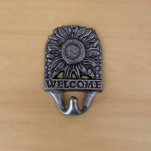 K&ouml;nnte beinhalten: Ein silberfarbener Metall-Wandhaken mit Sonnenblumen-Design und dem Wort "WELCOME". Der Haken hat ein Doppelhakendesign. Die Sonnenblume befindet sich in der Mitte des Hakens.