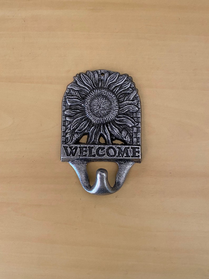 K&ouml;nnte beinhalten: Ein silberfarbener Metall-Wandhaken mit Sonnenblumen-Design und dem Wort "WELCOME". Der Haken hat ein Doppelhakendesign. Die Sonnenblume befindet sich in der Mitte des Hakens.