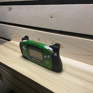 Puede incluir: Una Game Boy Advance SP verde con agarres negros. La pantalla es negra y los botones son blancos. El texto "GAME BOY ADVANCE" está impreso en la parte frontal del dispositivo.