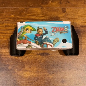 Puede incluir: Una consola de juegos portátil Nintendo DS blanca con una funda negra. La consola tiene un diseño personalizado con personajes del videojuego "The Legend of Zelda: Phantom Hourglass".
