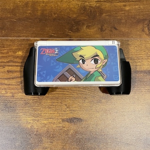 Puede incluir: Consola de juegos portátil Nintendo DS blanca con una funda protectora negra. La consola presenta una imagen colorida de Link del juego "The Legend of Zelda: Phantom Hourglass".