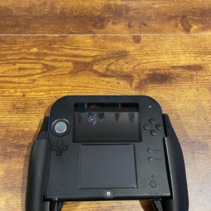 Può includere: Una console per videogiochi portatile Nintendo 3DS nera con uno stilo nero e grigio. La console ha uno schermo nero e un pad di controllo nero e grigio. La console è in una custodia nera con un'impugnatura nera e grigia.