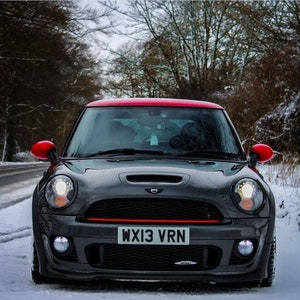 May include: A black Mini Cooper S with a red roof and red side mirrors. The car has a black license plate that reads "WX13 VRN". The car is parked in a snowy driveway.