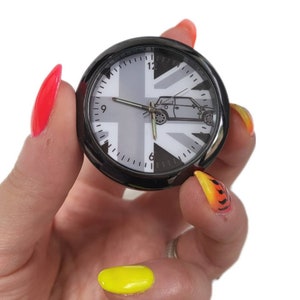 Può includere: Un orologio analogico nero e argento con un design Union Jack e una silhouette di un'auto. L'orologio è rotondo ed è tenuto in una mano con smalto per unghie dai colori vivaci.