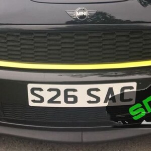 May include: A black Mini Cooper with a yellow stripe across the front bumper. The car has a black grille and a license plate that reads "S26 SAC".