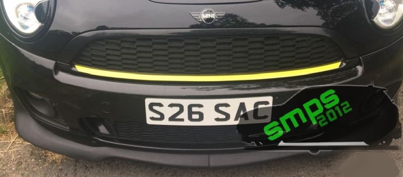 May include: A black Mini Cooper with a yellow stripe across the front bumper. The car has a black grille and a license plate that reads "S26 SAC".