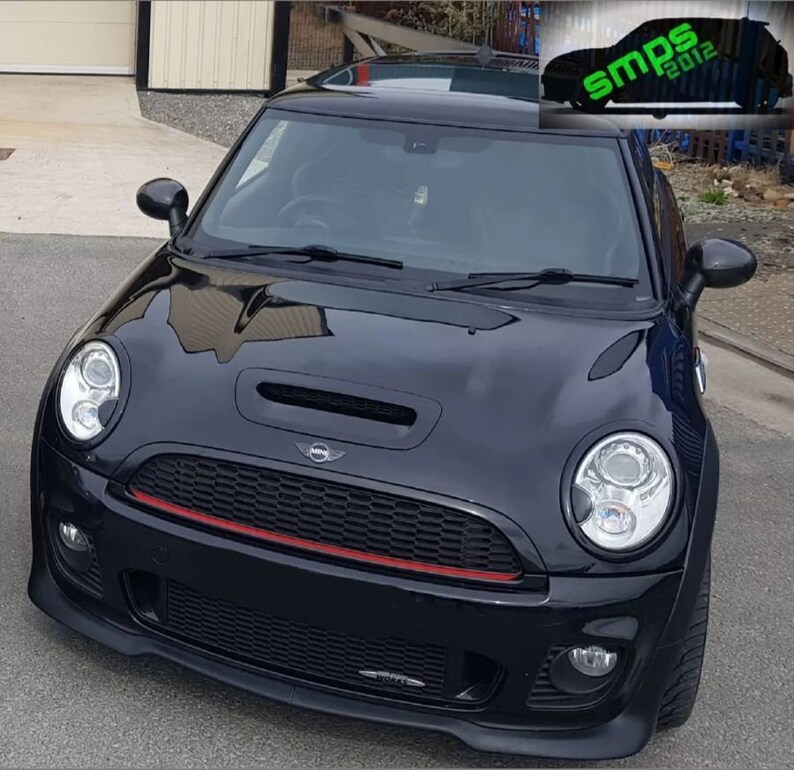 May include: A black Mini Cooper S with a red stripe on the front grille. The car has a black hood and a black bumper. The car is parked on a paved surface.