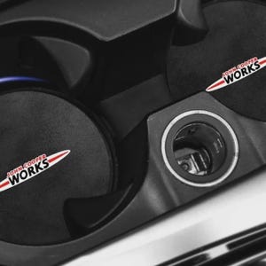 Pode incluir: Close-up do interior de um carro com porta-copos pretos e uma consola central. Os porta-copos têm bases redondas pretas com o logótipo "JOHN COOPER WORKS" em branco e vermelho. A consola central tem um anel prateado à volta de um pequeno compartimento.
