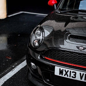 May include: A black Mini Cooper with a red stripe across the front bumper and a red side mirror. The car has a black grille with a chrome Mini logo and a license plate that reads "WX13 VI".