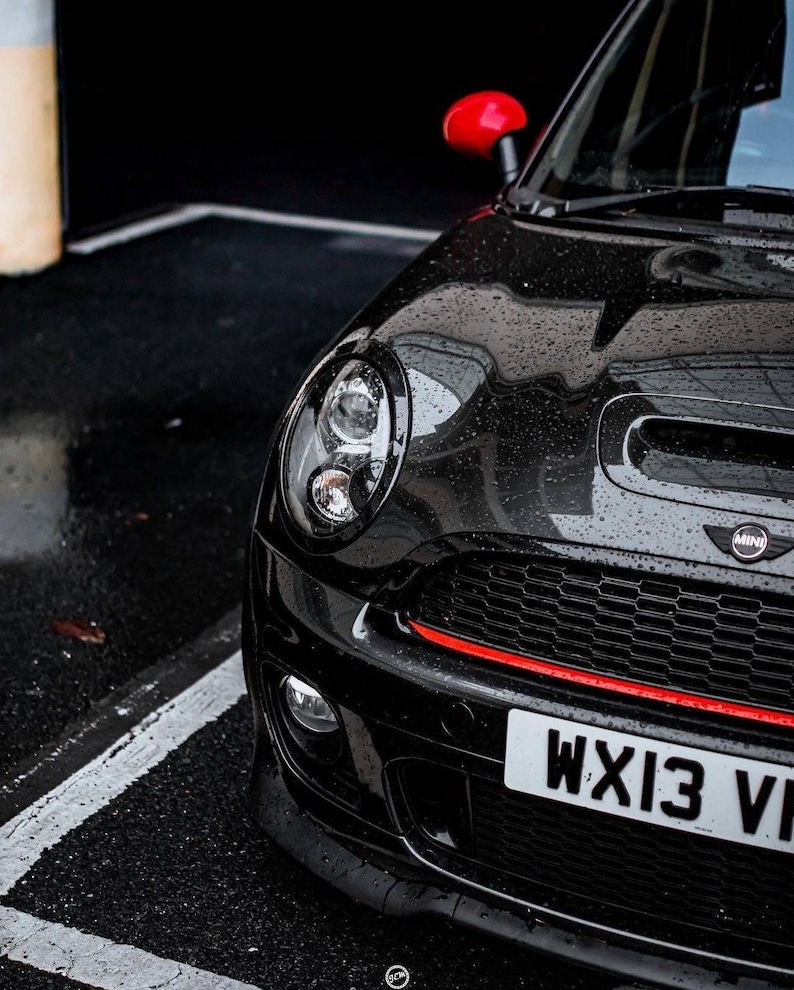 May include: A black Mini Cooper with a red stripe across the front bumper and a red side mirror. The car has a black grille with a chrome Mini logo and a license plate that reads "WX13 VI".
