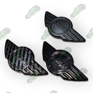 Puede incluir: Tres emblemas de coche negros con diseño de ala y la palabra "MINI" en un círculo. Uno tiene un patrón de fibra de carbono, los otros son negros lisos. Emblemas para un Mini Cooper.