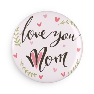 Könnte beinhalten: Ein runder, hellrosa Button mit den Worten "love you Mom" in handschriftlichem Stil. Das Wort "Mom" hat rosa Hasenohren. Kleine rosa Herzen und grüne Blätter verzieren den Button.