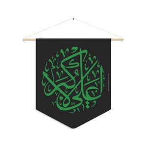 May include: A black banner with a green Arabic calligraphy design. The banner is suspended from a wooden rod with a string for hanging. The text on the banner is in a circular arrangement.