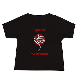 May include: Black t-shirt with red text and a white and red graphic. The text at the top reads "LABBAIK", and the text at the bottom reads "YA HUSSAIN". The graphic is a stylized Arabic script within a red circle.