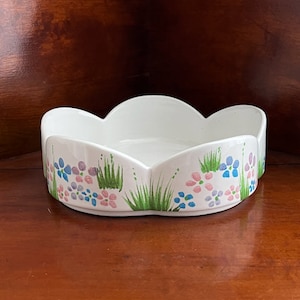 May include: White ceramic bowl with a scalloped edge, decorated with hand-painted pink, blue, and purple flowers, and green grass. The bowl is set on a wooden surface.