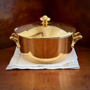 May include: A gold-coloured casserole dish with a lid, featuring decorative handles and a knob. It rests on a folded white napkin, reflecting the warm tones of the wooden surface. The design is classic and elegant, suitable for serving.